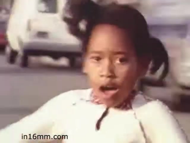 The still from 'Walk Safely - Educational Film from 1982 - Fiesta Films' features a young girl with pigtails, appearing startled or surprised, as she navigates a street setting. The background shows blurred vehicles, suggesting a busy environment. The frame likely emphasizes themes of safety and awareness in urban spaces, typical of educational films aimed at teaching children about traffic safety.