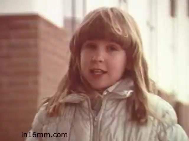 The still from 'Walk Safely,' an educational film from 1982 by Fiesta Films, features a young girl with light brown hair styled in a bob. She is wearing a puffy, light-colored jacket and appears to be speaking directly to the camera, likely delivering a safety message. The background shows a brick wall and some structure, suggesting an outdoor setting. The film appears to focus on pedestrian safety, aimed at educating children.