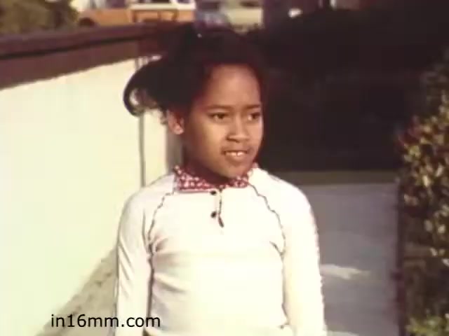 The still from the 1982 educational film 'Walk Safely' shows a young girl standing on a sidewalk. She appears to be wearing a white long-sleeve shirt with a decorative collar. Her hair is styled in a way that it falls loosely, giving her a youthful appearance. In the background, there is a fence and some greenery, suggesting a suburban setting. The overall vibe of the image is that of an educational moment, likely discussing pedestrian safety.