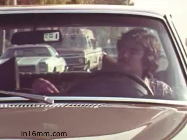 The still from 'Walk Safely' features a young man in the driver's seat of a vintage car. He appears relaxed, with his left arm resting on the steering wheel, and appears to be looking to his right. The background includes other vehicles, typical of a busy street scene. The film likely aims to convey safety messages, possibly highlighting road awareness during the early 1980s. The vintage aesthetic captures the style and context of the era effectively.