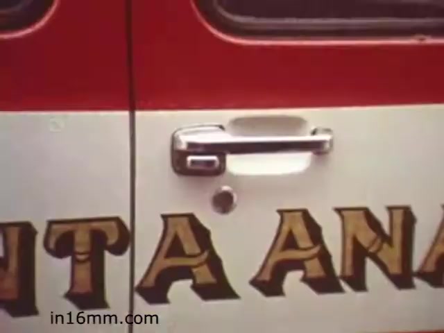 The still features a close-up of a vehicle door handle and part of the door panel, which is emblazoned with the words 'SANTA ANA' in bold, decorative lettering. The vehicle appears to be an older model, likely from the 1980s, consistent with the era of the film. The color scheme is predominantly red and white, typical of emergency vehicles or public service transport from that time. This scene likely serves to establish context for street safety or emergency response themes presented in the educational film.