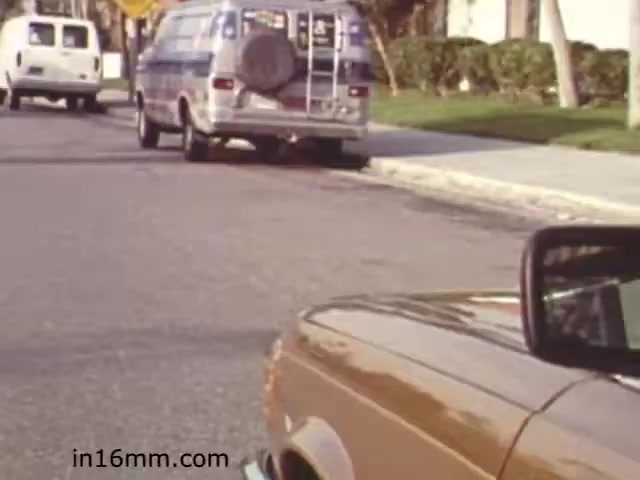 The still from 'Walk Safely,' an educational film from 1982, features a city street scene viewed from inside a car. In the foreground, part of the car's side mirror is visible, while the road stretches ahead. Two vehicles are parked nearby, one being a van with a spare tire mounted on its back. The setting appears suburban, with neatly trimmed grass and trees lining the street. This scene likely emphasizes pedestrian safety or awareness in a residential environment. The film is part of a series aimed at educating the public, possibly focusing on traffic safety and responsible behavior near roadways.
