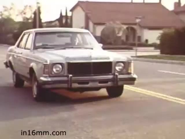The still from 'Walk Safely' (1982) features a vintage car driving down a suburban street. The setting looks typical of the era, with a residential home in the background and trees lining the road. The vehicle appears to be a mid-sized sedan, indicative of the 1970s or early 1980s design. The overall scene likely aims to illustrate safe walking practices in a community setting, consistent with the educational nature of the film.