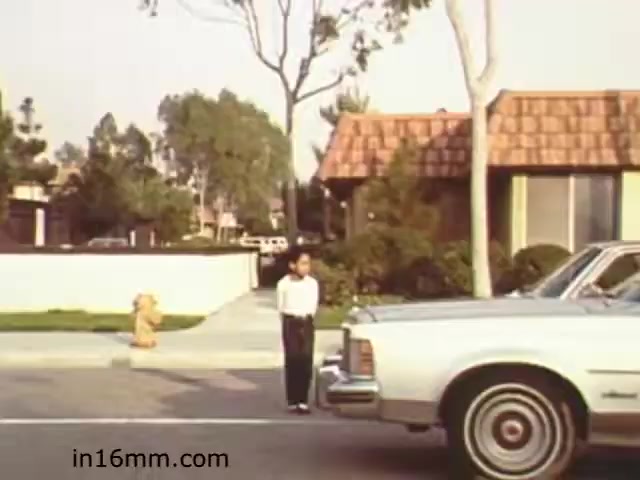 The 16mm still from 'Walk Safely,' an educational film from 1982 by Fiesta Films, depicts a child standing on a sidewalk, seemingly waiting to cross the street. In the foreground, a classic car is parked on the road. The background features suburban homes with well-maintained lawns and a fire hydrant visible on the left. The scene captures a moment emphasizing pedestrian awareness and safety in a residential neighborhood.