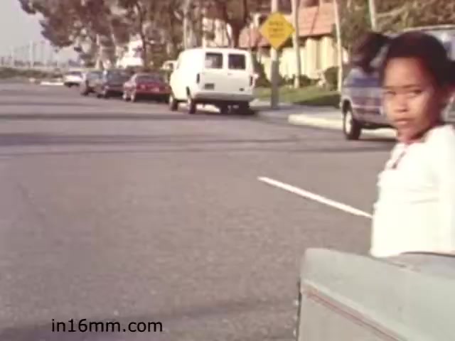 The still from the 1982 educational film 'Walk Safely' depicts a street scene with a young girl looking back towards the viewer, standing near a parked car. The background includes several vehicles and a street sign, emphasizing a residential environment. The film likely focused on teaching safety awareness, particularly for children, in pedestrian contexts. The vintage aesthetic reflects the period's style and approach to educational content.