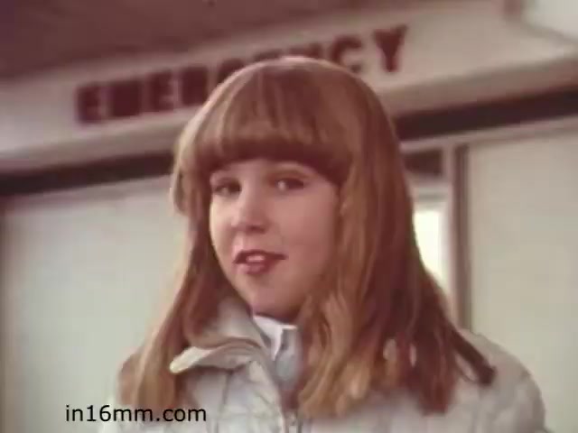 The still from 'Walk Safely,' an educational film from 1982, features a young girl with a distinctive blonde bob haircut, wearing a light jacket. She appears to be standing outside what looks like an emergency room, indicated by the sign in the background. The tone seems to convey a friendly, approachable vibe, likely as she prepares to convey a safety message. The aesthetic reflects the style of the early '80s educational films, which often aimed to instruct youth on various topics, including safety.
