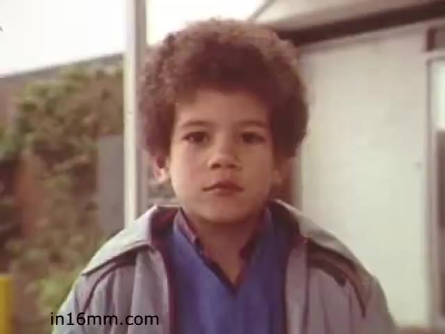 The still from the educational film 'Walk Safely' features a young boy with curly hair, wearing a light jacket over a blue shirt. He has a serious expression, possibly conveying a sense of focus or determination. The background hints at an outdoor setting, with soft natural lighting. This still likely relates to themes of safety and awareness, consistent with the film's educational purpose.