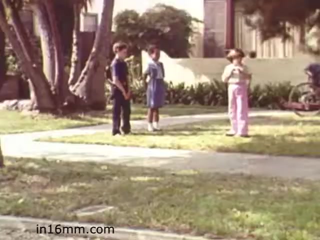 The still from 'Walk Safely,' an educational film from 1982, features three children standing on a grassy area near a sidewalk. The boys are wearing casual clothing, while one girl appears to be holding something in her hands. The setting seems to be a suburban neighborhood, with lush greenery and some residential buildings in the background. The children's expressions suggest they might be engaged in a discussion or activity related to safety. The film likely aims to teach safety awareness in a playful, relatable manner for children.
