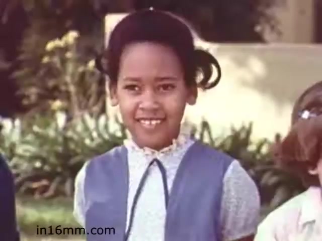 The still from 'Walk Safely,' produced by Fiesta Films in 1982, features a young girl smiling, dressed in a blue outfit. Her hair is styled with playful curls. The background shows greenery, suggesting an outdoor setting. The film likely focuses on safety education, highlighting themes of community and child awareness.