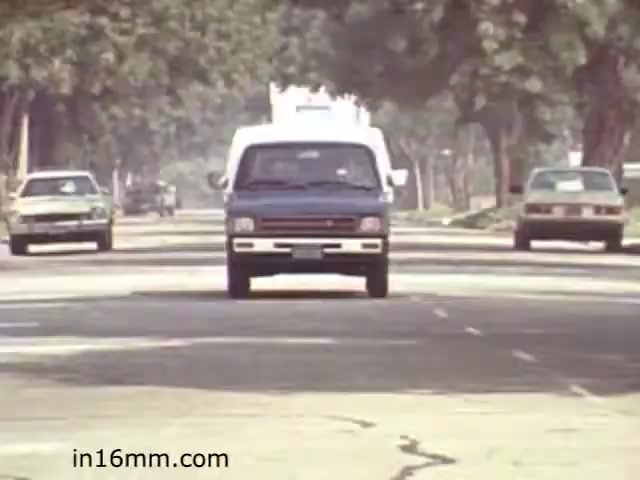 The still from 'Walk Safely,' an educational film from 1982, depicts a street scene with vehicles driving along a tree-lined road. In the foreground, a blue van is prominently featured, suggesting an emphasis on traffic safety. Other cars are visible in the background, contributing to an urban setting. The overall composition likely aims to convey lessons about pedestrian safety in an everyday environment.
