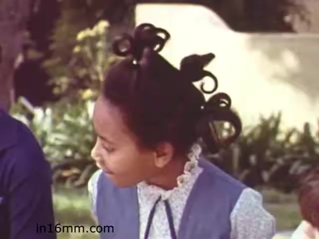 The still from 'Walk Safely,' an educational film from 1982, features a young girl with distinctive hair styled in multiple curls, wearing a blue dress with a white collar. She appears engaged in conversation, possibly with peers, in a vibrant outdoor setting that includes greenery. The film likely focuses on childhood safety themes, emphasized by the children's attire and expressions.