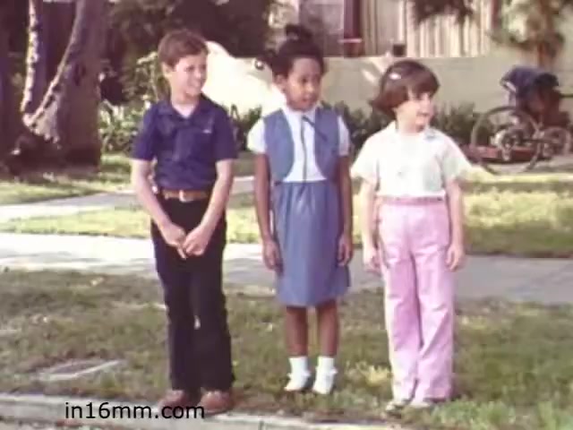 The still from 'Walk Safely,' an educational film from 1982 by Fiesta Films, features three children standing together on a sidewalk. They appear to be in a suburban setting, with grass and a house in the background. The boy on the left is wearing a blue shirt, black pants, and brown shoes, while the girl in the middle is dressed in a blue dress with a white collar, and the girl on the right is wearing a light pink shirt and pink pants. Their expressions and posture suggest they are engaged in a lesson about safety, likely related to walking and being aware of their surroundings. The film aims to educate children on safety practices.