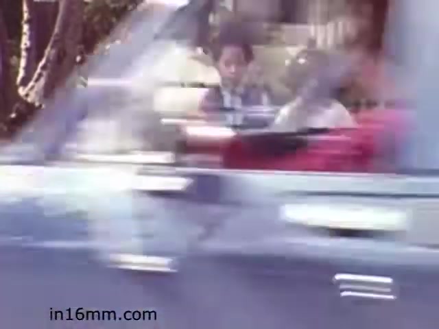 The still from 'Walk Safely,' an educational film from 1982, captures a moment where children appear to be in a car as it drives by. The focus is somewhat blurred, suggesting motion, possibly to emphasize the importance of being aware of traffic while walking. The film likely aims to communicate safety tips for pedestrians, particularly for kids. The context of the scene reflects the educational theme of promoting road safety awareness.