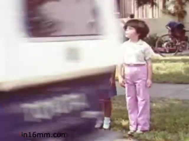 The still from 'Walk Safely,' an educational film from 1982 by Fiesta Films, features a young child dressed in a light shirt and pink pants standing on a sidewalk. In the background, a vehicle is seen moving past, emphasizing a safety message regarding pedestrian awareness. Another child is partially visible behind the main subject, hinting at a communal context. The scene suggests a focus on teaching children about road safety and the importance of being cautious around traffic. The overall tone is likely educational and informative, typical of films from that era aimed at children.