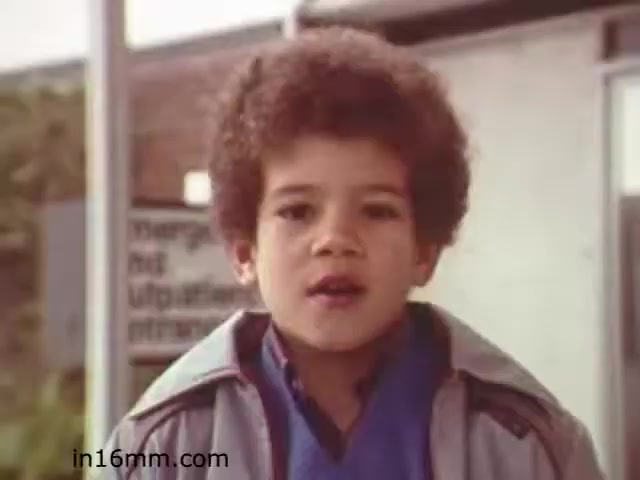 The still from 'Walk Safely,' an educational film from 1982, features a young boy with curly hair speaking directly to the camera. He appears to be outdoors, with a background that suggests a school or community environment. The film likely focuses on pedestrian safety, given its title and educational purpose. The boy's expression is earnest, reflecting the seriousness of the message. Overall, the image captures a moment intended to engage and inform viewers about safety practices.
