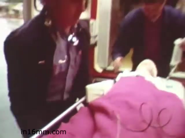 The still from 'Walk Safely - Educational Film from 1982' depicts a scene inside an ambulance. Two individuals, likely emergency responders, are seen assisting a patient, who is covered with a blanket. The atmosphere suggests a focus on emergency medical response and safety, which aligns with the film's educational theme about walking safely and awareness in emergency situations. The overall tone appears serious, highlighting the importance of medical assistance.
