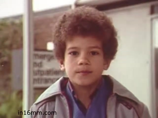 The still from 'Walk Safely' features a young boy with curly hair, dressed in a blue shirt and a light jacket. He appears to be in an educational environment, possibly a school setting. The background suggests a focus on safety awareness, although specific details about the surroundings are minimal. The overall tone of the film is likely aimed at teaching children about safety protocols, particularly in relation to walking and traffic awareness.