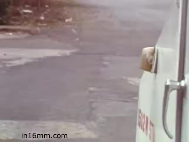 The still from 'Walk Safely' appears to show a close-up of a vehicle parked at a somewhat deserted intersection or street. The vehicle, possibly a truck or service van, is partially in the frame, with its side visible. The background features a blurry street scene that seems quiet, with asphalt visible and some debris or grass at the edges of the road. The overall tone suggests an emphasis on safety while walking, likely reflecting the educational objective of the film from the early 1980s.