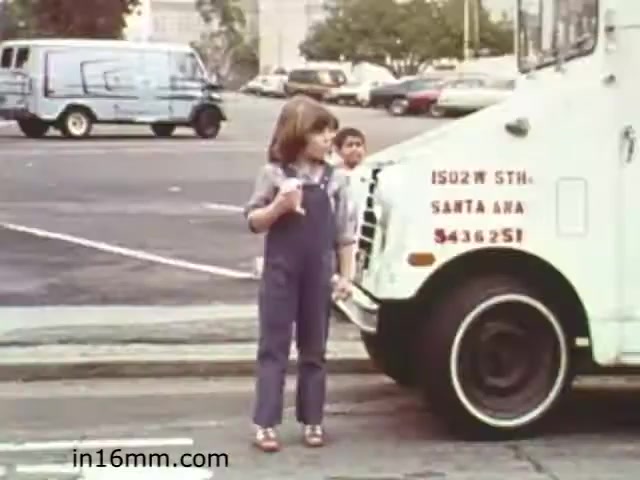 The still from 'Walk Safely,' an educational film from 1982, depicts a young girl in overalls standing near the front of a white truck. She appears to be holding something (possibly a sandwich or snack) and is looking downward, while a boy peeks out from behind the truck. The scene captures a parking lot setting with several vehicles in the background, suggesting a focus on safety in pedestrian environments. The film likely aims to educate children on being cautious around vehicles.