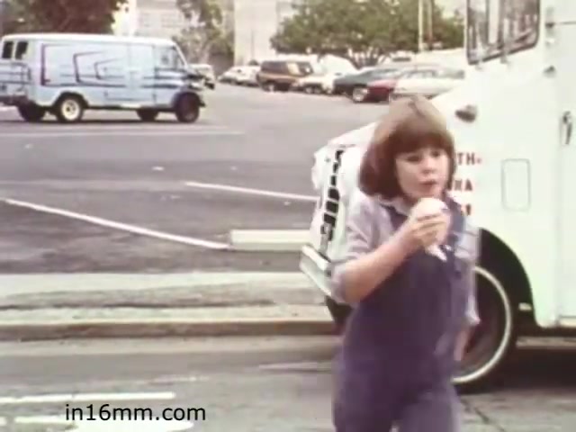 The still from 'Walk Safely,' an educational film from 1982 by Fiesta Films, features a young girl walking in a parking lot while enjoying an ice cream cone. She is dressed in a casual outfit, with light denim overalls. In the background, vehicles are parked, and there is a food truck nearby, possibly selling ice cream. The scene captures a moment that likely emphasizes pedestrian safety, showcasing a typical social setting from that era.