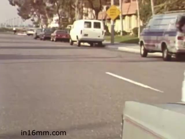 The still from 'Walk Safely,' an educational film from 1982, shows a suburban street scene. In the foreground, there is a vehicle parked on the right, and several other cars are visible along the street. To the left, a white van and another vehicle are parked. A sign indicating a crosswalk or pedestrian caution is positioned alongside the road, suggesting the film's focus on pedestrian safety. The image conveys a typical residential area, likely used to illustrate safe practices for walking in such environments.