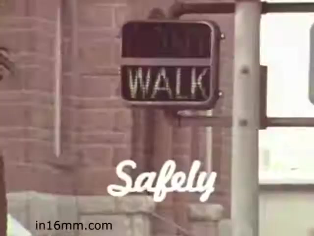The still from 'Walk Safely,' an educational film from 1982 by Fiesta Films, features a pedestrian traffic signal displaying the word 'WALK.' The sign is framed against a backdrop that includes a brick wall and part of a traffic pole. The film likely aims to promote pedestrian safety, implying that it’s safe for pedestrians to cross the street. The aesthetic reflects the style of the early '80s, contributing to its educational theme.