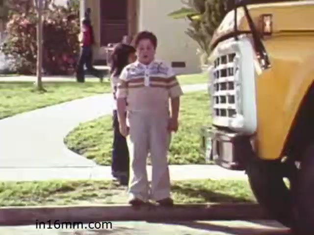 The still from 'Walk Safely,' an educational film from 1982, depicts two children near a yellow school bus. One boy, wearing a striped shirt and light-colored pants, stands prominently in the foreground, appearing startled or concerned. A girl is seen in the background, positioned further along the sidewalk. The scene is set on an urban street with grassy areas, suggesting a typical neighborhood. The film likely focuses on pedestrian safety, emphasizing awareness around school buses.