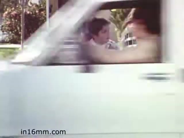 The still from the film 'Walk Safely' shows a scene depicting a moving vehicle, suggesting a moment focused on road safety. The image captures two individuals in the car, with one appearing to look toward the viewer. The background features greenery, typical of suburban or park-like settings. The overall tone aims to convey themes of safety and awareness in a casual, educational context suitable for its 1982 release.