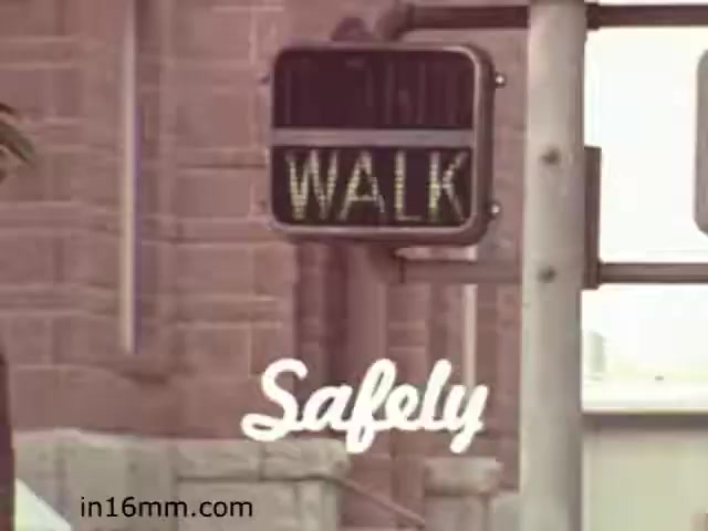 The still from the 1982 educational film 'Walk Safely' features a pedestrian traffic signal displaying the word 'WALK' in illuminated letters. Below the signal, the word 'Safely' is artistically rendered in a cursive font. The background suggests an urban environment, characterized by buildings and possibly a tree or foliage. Overall, the image emphasizes pedestrian safety and awareness, aligning with the film's educational purpose.