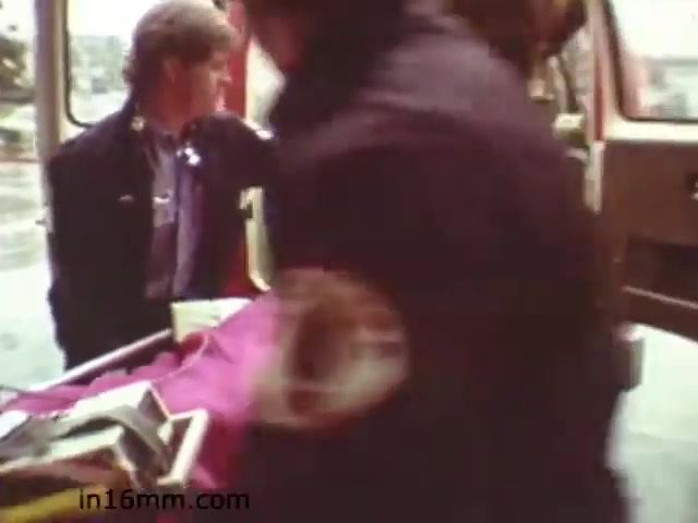 The still from 'Walk Safely,' an educational film from 1982, appears to depict a scene involving emergency responders. It shows a person seated in an ambulance, while paramedics are in the process of transporting a patient on a stretcher. The atmosphere suggests urgency, highlighting the importance of safety and preparedness in emergency situations. The overall aesthetic reflects the filmmaking style of the early 1980s.
