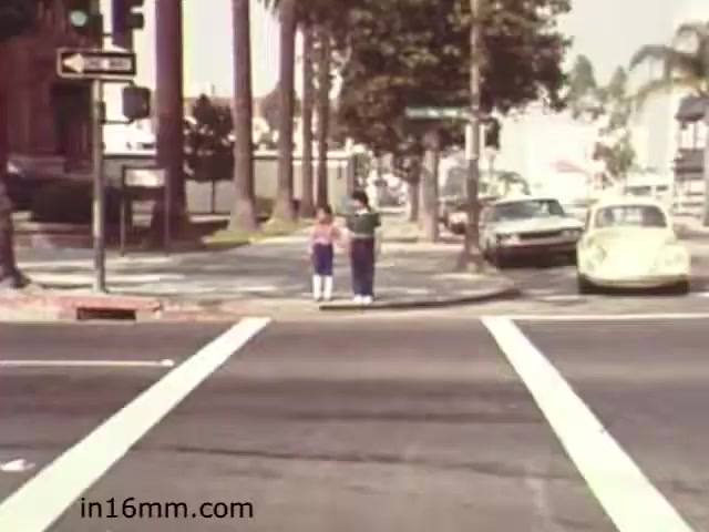 The still from 'Walk Safely - Educational Film from 1982' shows two children standing at a crosswalk, preparing to cross the street. The scene is set in a suburban area with palm trees lining the street, indicative of a warm climate. Traffic signals and parked cars, including a classic Volkswagen Beetle, add to the context of urban safety education. This film likely focuses on teaching children how to navigate road safety and pedestrian awareness. The overall presentation is reflective of educational materials from the early 1980s.