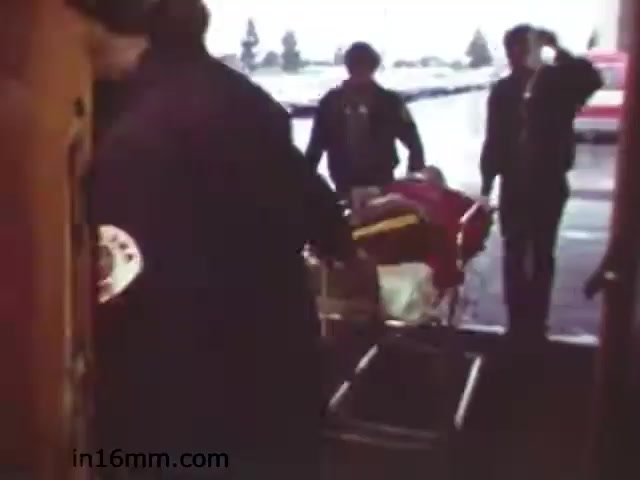 The still from 'Walk Safely - Educational Film' appears to show a scene focusing on safety or emergency response. It depicts individuals, including emergency personnel, handling a stretcher, suggesting a situation requiring urgent care or assistance. The setting seems to be outside, likely near a vehicle, possibly an ambulance. The atmosphere is serious, reflecting the importance of safety protocols. The film likely aimed to educate viewers on emergency procedures and the importance of safety awareness.