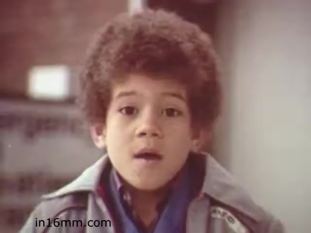The still depicts a young boy with curly hair, wearing a light-colored jacket over a blue shirt. He appears to be speaking or engaging with the audience, likely conveying a message related to safety. The overall tone suggests a serious yet approachable educational theme, typical of children's safety films from the early 1980s. The film 'Walk Safely' is likely aimed at teaching children about pedestrian safety and awareness.