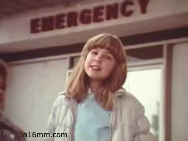 The still from 'Walk Safely,' an educational film from 1982, features a young girl with medium-length, straight hair, speaking directly to the camera. She appears to be offering a message or advice related to safety. In the background, there's a sign that reads 'EMERGENCY,' suggesting a context focused on health or safety awareness. The overall tone of the image suggests an educational atmosphere aimed at teaching young audiences about safety practices.
