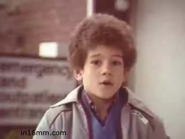 The still from 'Walk Safely,' an educational film from 1982, features a young boy with curly hair speaking directly to the camera. He appears to be outdoors, with a sign in the background that suggests a focus on safety or guidance, typical of educational content aimed at children. The film likely addresses pedestrian safety and awareness, presented through friendly, relatable characters to engage its audience. The boy's expression suggests he is eager to convey important information.