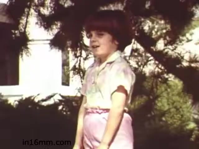 The still from 'Walk Safely,' an educational film from 1982 by Fiesta Films, features a young child standing outdoors, likely in a school or community setting. The child appears to be wearing a light-colored shirt and pink pants. Surrounding foliage suggests a focus on safety in outdoor environments. The film likely aims to educate viewers on safety practices, possibly targeting children and their interactions with pedestrian spaces.
