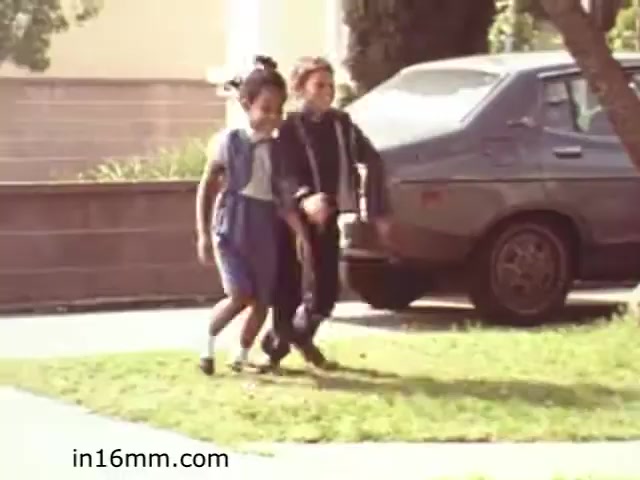 The still from 'Walk Safely,' an educational film from 1982 by Fiesta Films, depicts two children walking together on a grassy area, presumably while learning about pedestrian safety. One child wears a blue outfit with a vest, while the other is dressed in a black jacket. In the background, a parked car is visible, suggesting a suburban or neighborhood setting. This scene likely emphasizes the importance of walking safely in areas where vehicles are present.