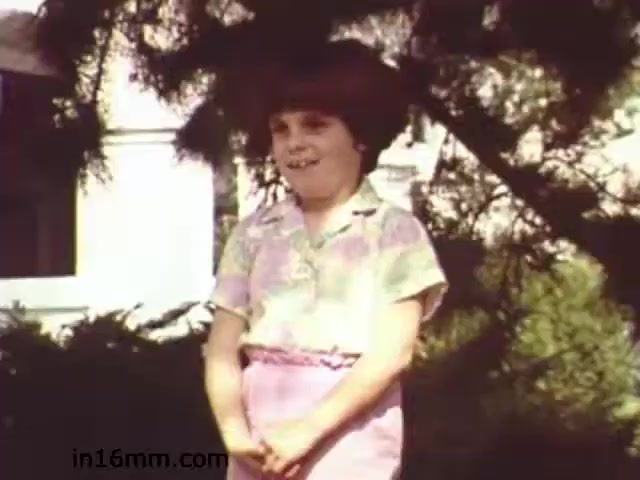 The still from 'Walk Safely,' an educational film from 1982 by Fiesta Films, features a young girl with a cheerful expression, standing outside. She wears a colorful, short-sleeved shirt and a pink skirt. The background includes greenery and what appears to be a building, suggesting an outdoor setting. The image captures a youthful energy, typical of educational films aimed at teaching safe walking practices.