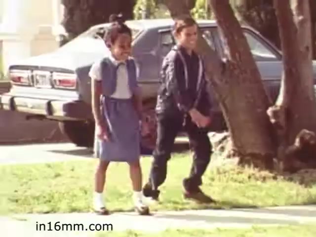The still from 'Walk Safely,' an educational film from 1982, features two children walking along a sidewalk. One child is wearing a navy blue dress and white socks, while the other is in a black jacket and pants. They appear to be engaged in conversation as they walk, with a car parked in the background, suggesting a suburban setting. The image reflects a focus on pedestrian safety for children, typical of educational films from that era.