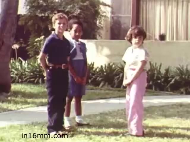 The still from 'Walk Safely,' an educational film from 1982, showcases three children standing together on a sidewalk in a residential area. The children are dressed in casual summer clothing, with the background featuring greenery and a house. The scene likely emphasizes themes of safety and community, consistent with the educational objectives of the film. The lighting and color palette reflect the typical aesthetic of the early '80s, contributing to its nostalgic feel.