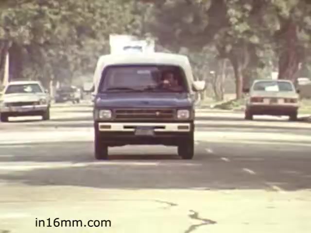 The still from 'Walk Safely,' an educational film from 1982 by Fiesta Films, features a blue van driving down a tree-lined street. The scene suggests a focus on road safety, likely showcasing typical traffic conditions of the time. The vehicles in the background indicate a suburban or city environment, emphasizing the importance of pedestrian awareness. The vintage aesthetic reflects the era of the film, contributing to its educational context.