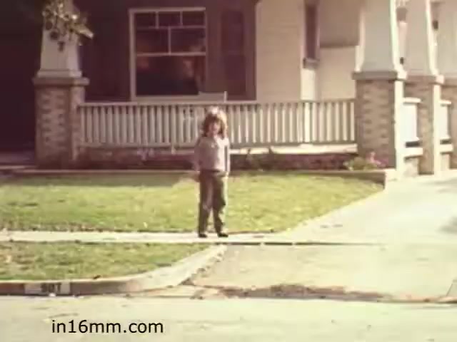 The still from 'Walk Safely,' an educational film from 1982, features a young child standing on a sidewalk in front of a residential house. The child appears to be observing the street, suggesting a theme related to pedestrian safety. The environment includes well-kept lawns and a porch, typical of suburban settings. The image conveys a sense of innocence and the importance of being cautious while walking near traffic.