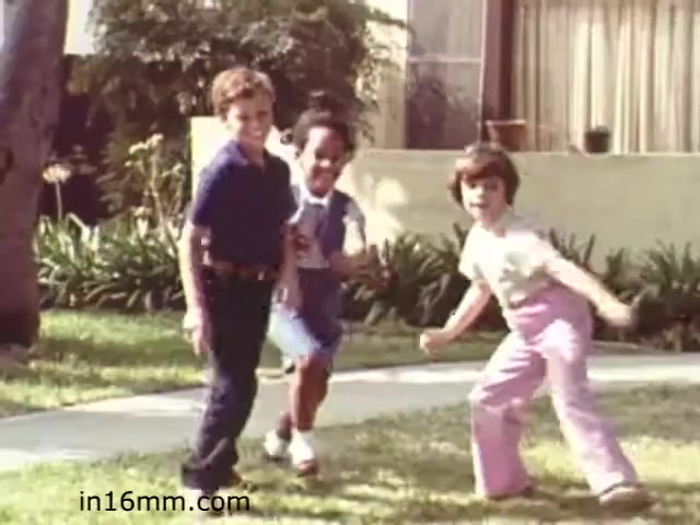 The still from 'Walk Safely,' an educational film from 1982 by Fiesta Films, shows three children playing in a grassy yard. They appear to be having fun, with smiles on their faces. The setting features a suburban backdrop with a house in the background and vibrant greenery. The children's attire reflects the fashion of the early 1980s, showcasing a casual and playful atmosphere typical of childhood. This film likely aimed to promote safety awareness in a relatable and engaging manner for kids.