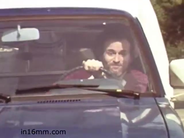 The still from the 1982 educational film 'Walk Safely' shows a man driving a vehicle, likely part of a safety message related to pedestrian awareness or road safety. His expression appears cheerful, which may suggest a lighthearted or engaging approach to the serious topic. The setting seems casual, indicative of the film's educational style aimed at making safety relatable to viewers. This film likely emphasizes the importance of being aware of one's surroundings while driving and walking.
