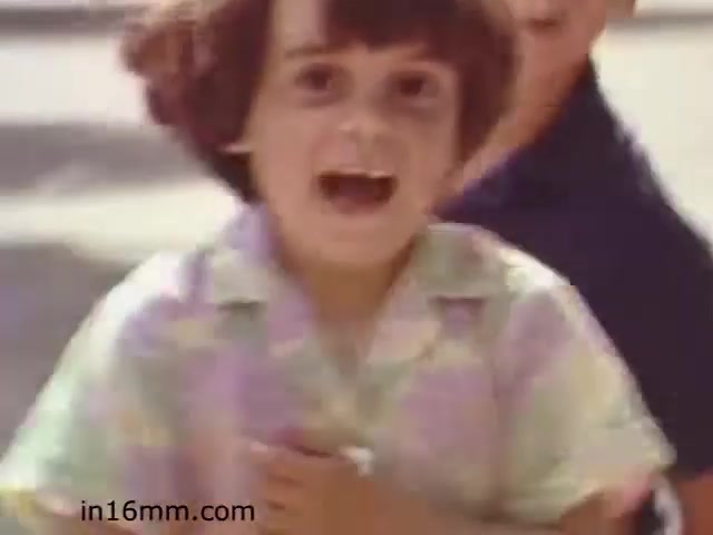 The still from 'Walk Safely,' an educational film from 1982, features a young child with a bright expression, possibly running or playing outdoors. The child is wearing a colorful, patterned shirt and is engaging joyfully with others in the background, suggesting a light-hearted theme. The film likely focuses on safety awareness for children, with a positive, energetic vibe that encourages interaction and play.