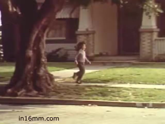 The still from 'Walk Safely,' an educational film from 1982 by Fiesta Films, depicts a young child running on a well-maintained sidewalk in a suburban neighborhood. In the background, there is a large tree and a house, suggesting a residential setting. The scene likely emphasizes themes of safety while walking, typical of educational materials from that era aimed at teaching children about pedestrian safety. The overall tone is nostalgic, capturing a simpler time in suburban life.