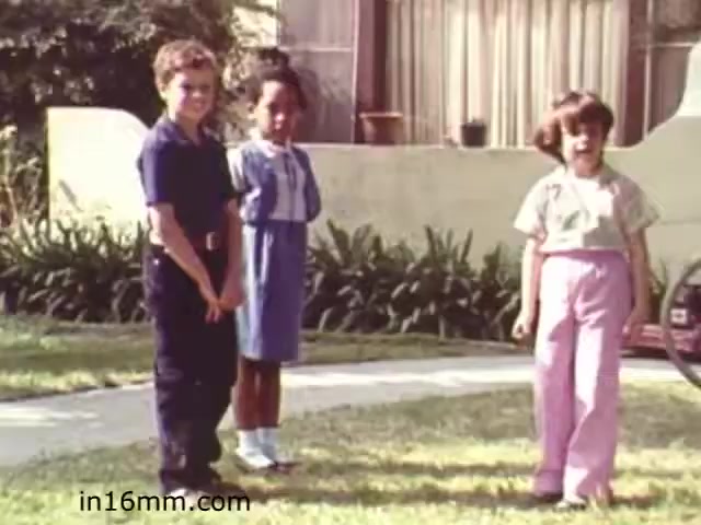 The still from 'Walk Safely,' an educational film from 1982, features three children standing on a grassy lawn. Two boys and a girl are positioned in a casual manner, wearing colorful clothing typical of the era. The background includes a single-story house with large windows and greenery, suggesting a suburban setting. The film likely focuses on pedestrian safety, promoting awareness among children in a relatable, engaging way.