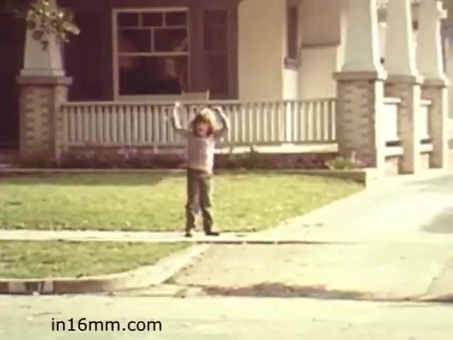 The still from 'Walk Safely,' an educational film from 1982, features a child standing on a sidewalk, appearing to signal or draw attention, against the backdrop of a suburban house with a porch. The scene captures a typical neighborhood setting, likely emphasizing safety while walking or playing in the area. The film aims to educate viewers about pedestrian safety, reflecting the era's concerns for children and their well-being in public spaces.