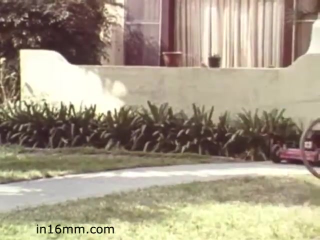 The still from 'Walk Safely,' an educational film from 1982 by Fiesta Films, features a suburban setting with a pathway lined by a neatly maintained lawn. In the foreground, a bicycle is partially visible, suggesting a focus on pedestrian safety and possibly bicycle awareness. The backdrop consists of a residential house with large windows and a decorative plant by the entrance. The overall scene conveys a sense of community safety, likely aimed at educating viewers on safe practices in neighborhood environments.