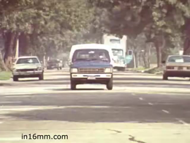 The still from 'Walk Safely,' an educational film from 1982 by Fiesta Films, depicts a suburban street scene. It features a mix of vehicles, including a prominent blue van driving towards the camera, surrounded by other cars and a truck in the background. The setting has a tree-lined roadway, indicative of a residential area, with visible pavement and some wear. The scene likely aims to convey themes related to road safety and awareness for pedestrians and drivers.